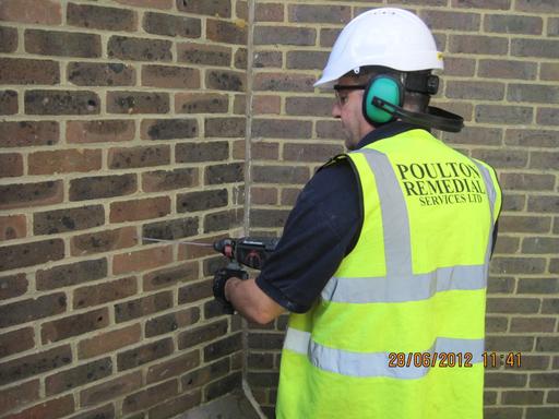 Poulton Remedial worker wearing Hi-Viz vest, hardhat and hearing protectors whilst drilling into a wall